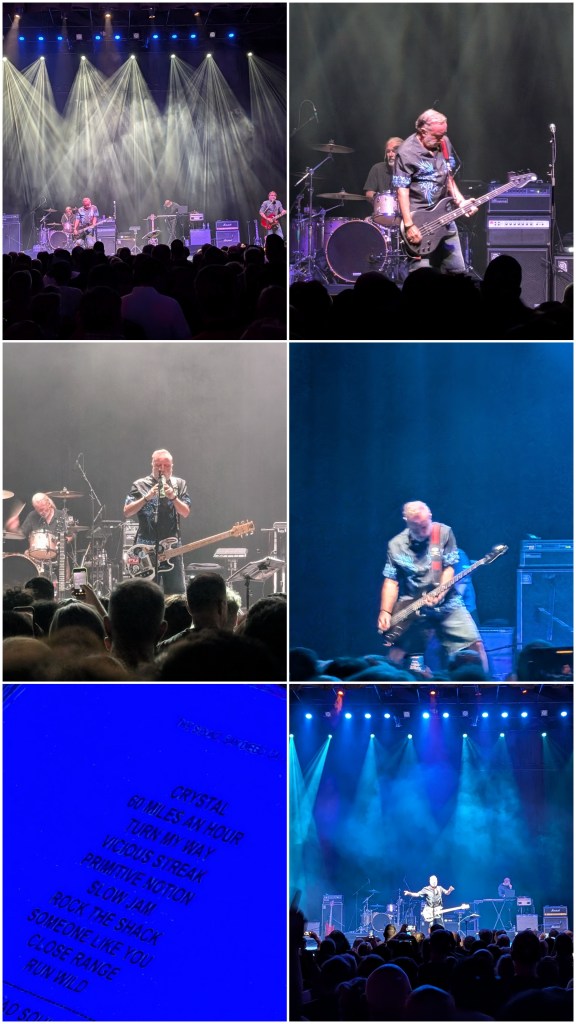 A live music performance with a band on stage under dramatic lighting, crowd visible in front.
