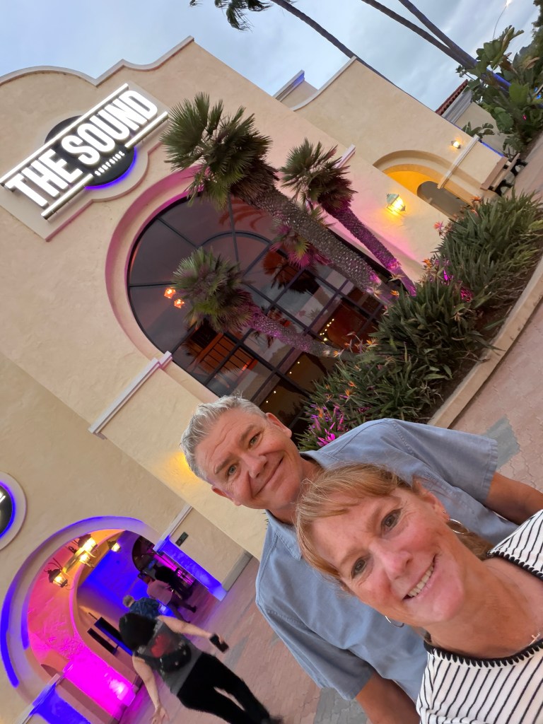 A couple poses for a selfie in front of a building with a sign that reads 'THE SOUND.' The exterior is adorned with tropical plants, and the scene is illuminated with colorful lights.