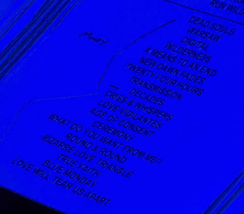 A list of song titles in blue lighting, featuring tracks like 'Dead Souls', 'Love Will Tear Us Apart', and 'Blue Monday'. The text has a neon effect against a dark background.